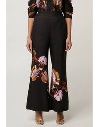A model wearing black wide-leg pants with a large brown, orange, and pink floral print on the lower legs, paired with a matching patterned top and beige sandals, against a light gray studio background.