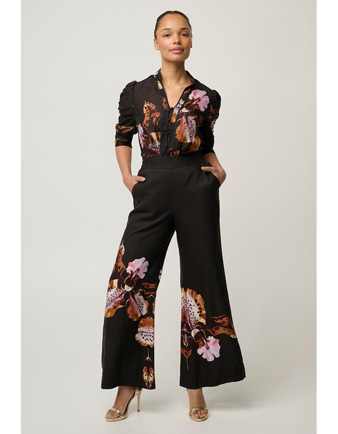 A model wearing a black wide-leg pants jumpsuit with a floral print in pink, orange, and brown, paired with a matching black and floral long-sleeve top, against a light gray studio background.