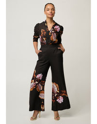 A model wearing a black wide-leg pants jumpsuit with a floral print in pink, orange, and brown, paired with a matching black and floral long-sleeve top, against a light gray studio background.