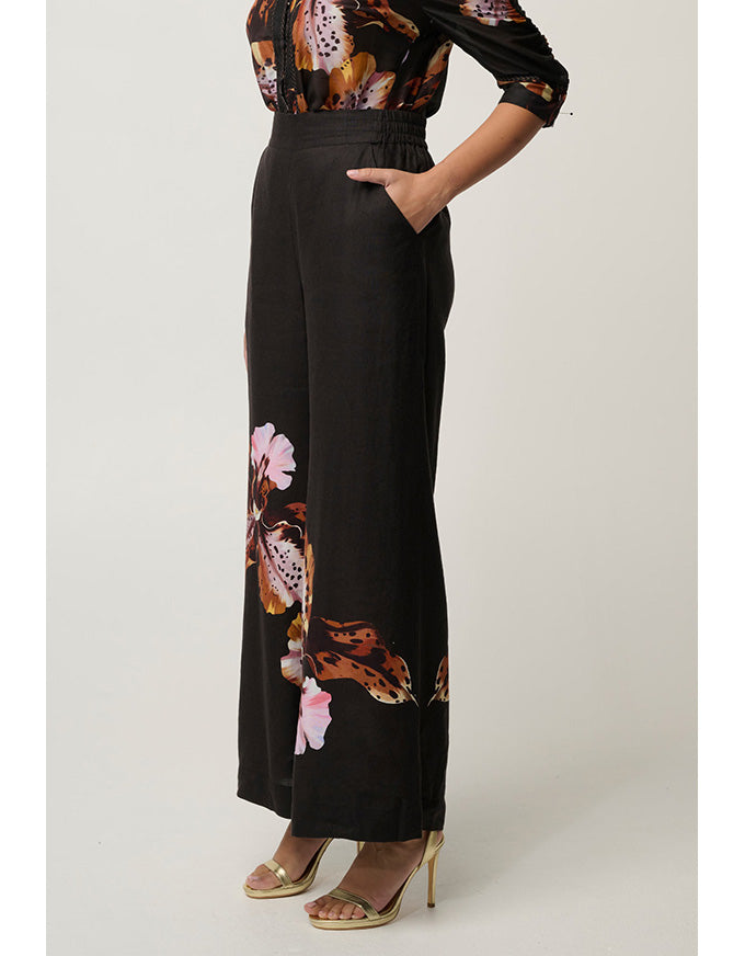 A model is wearing a black wide-leg jumpsuit with a brown, pink, and white floral print along the side and hem, paired with gold high-heeled sandals against a light gray studio background.