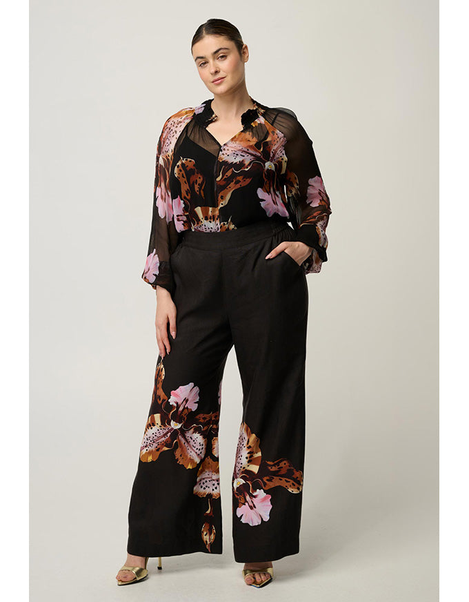 A woman models a black wide-leg pants set with a matching sheer long-sleeve top featuring a brown, pink, and orange floral print, photographed against a neutral light gray studio background.