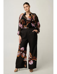 A woman models a black wide-leg pants set with a matching sheer long-sleeve top featuring a brown, pink, and orange floral print, photographed against a neutral light gray studio background.