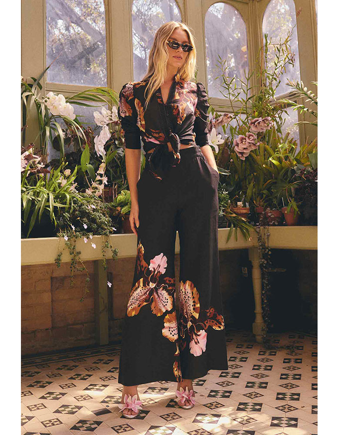 A woman models a black two-piece jumpsuit with a floral print in a sunlit greenhouse. The jumpsuit features a short-sleeve button-up top with a tie waist and matching wide-leg pants with large floral details near the hem.