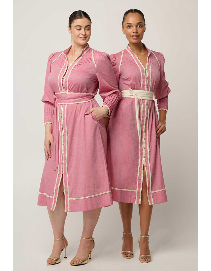 Two women modeling matching pink long-sleeve midi dresses with cream trim and self-tie belts, standing against a plain light beige studio background.