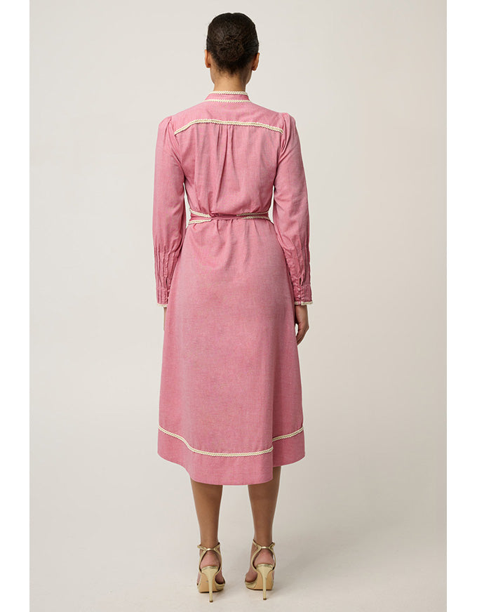 A model viewed from the back wearing a pink long-sleeve midi dress with cream-colored piping details, paired with beige high-heeled sandals, against a light beige studio background.