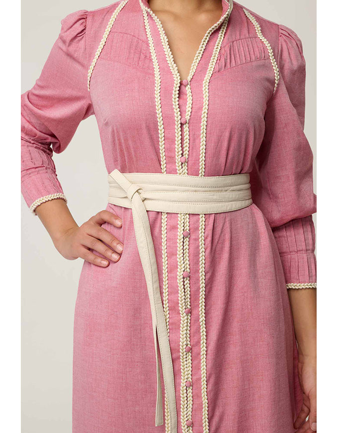 A pink long-sleeve dress with cream-colored braided trim along the front opening, collar, cuffs, and waist, featuring a matching cream tie belt at the waist.