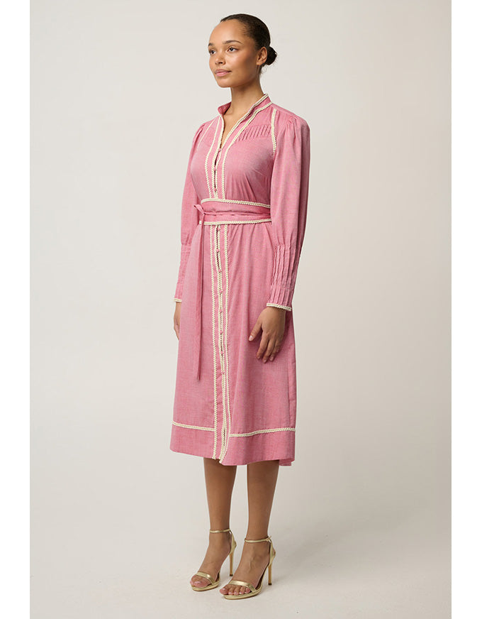 A woman modeling a pink long-sleeve midi dress with cream-colored trim and a matching waist tie, photographed against a neutral light gray studio background.
