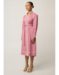 A woman modeling a pink long-sleeve midi dress with cream-colored trim and a matching waist tie, photographed against a neutral light gray studio background.