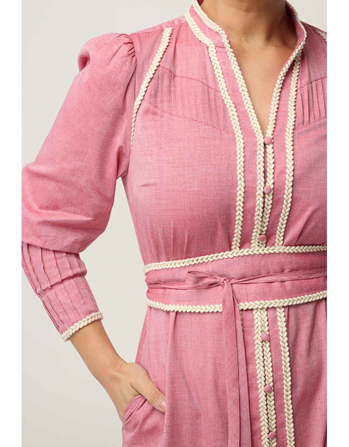 A close-up of a pink long-sleeve dress with cream braided trim along the neckline, front opening, cuffs, and waist tie, shown on a model against a light neutral background.