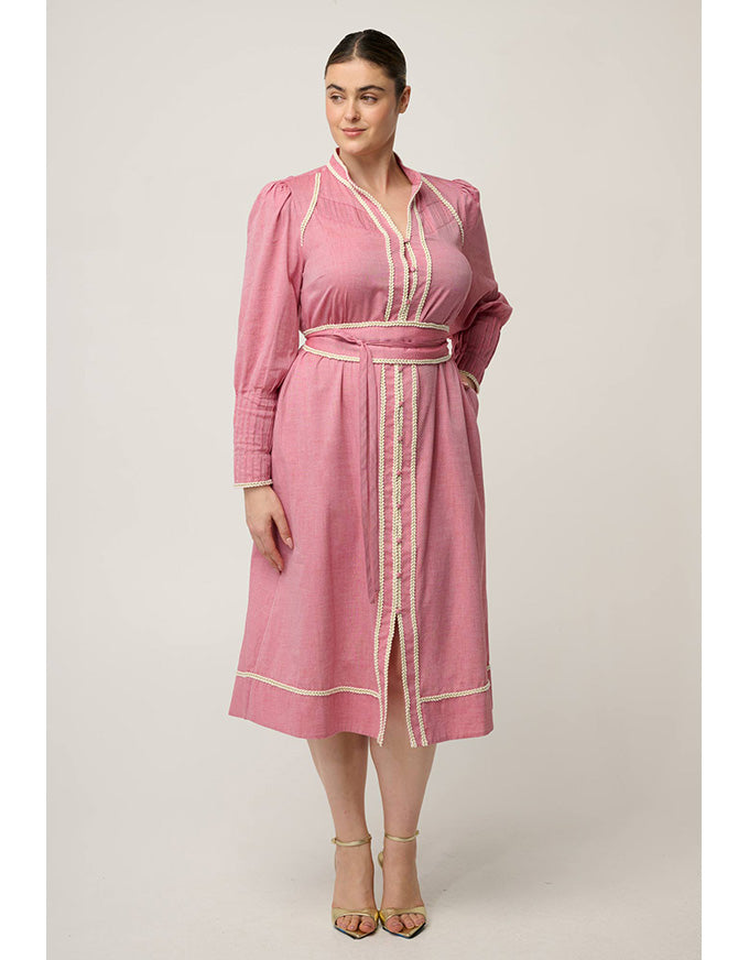 A woman modeling a pink long-sleeve midi dress with cream-colored trim and a matching belt, standing against a neutral light gray studio background.