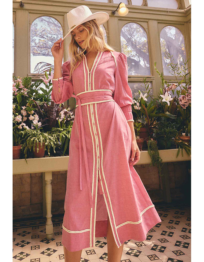 A woman wearing a pink midi dress with cream trim and a matching hat, standing in a sunlit greenhouse with plants and arched windows in the background.