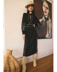Woman in a black dress with white trim, beige boots, and hat standing in a room with a painting on the wall.