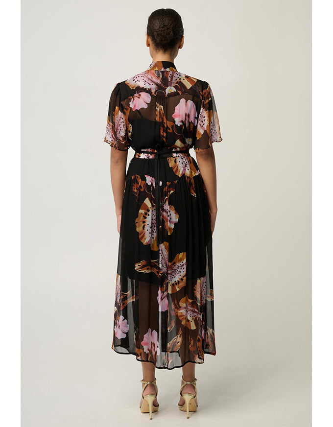 A woman viewed from the back wearing a sheer black midi dress with a pink, brown, and beige floral print, tied at the waist with a matching belt, paired with beige high-heeled sandals against a light gray studio background.