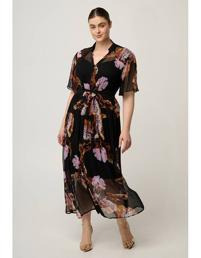 A woman modeling a black midi dress with a sheer overlay and a brown, pink, and beige floral print. The dress features short sleeves, a V-neckline, and a matching fabric tie belt at the waist, shown against a plain light gray studio background.