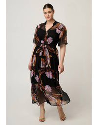 A woman modeling a black midi dress with a sheer overlay and a brown, pink, and beige floral print. The dress features short sleeves, a V-neckline, and a matching fabric tie belt at the waist, shown against a plain light gray studio background.