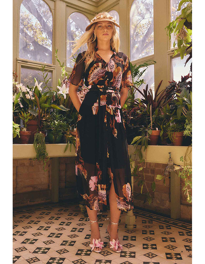 A woman stands in a sunlit greenhouse wearing a sheer black midi dress with a pink, orange, and beige floral print, a matching hat, and pink shoes. The background features potted plants and large arched windows.