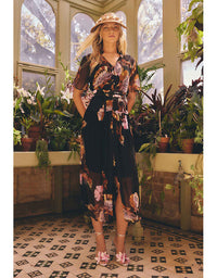 A woman stands in a sunlit greenhouse wearing a sheer black midi dress with a pink, orange, and beige floral print, a matching hat, and pink shoes. The background features potted plants and large arched windows.