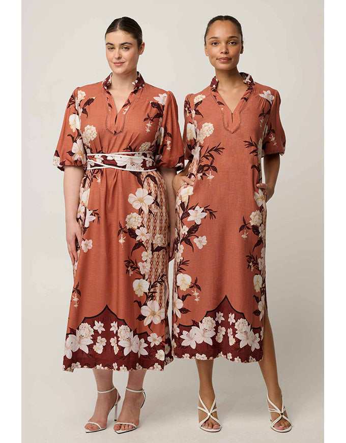 Two models wearing matching rust-orange floral midi dresses with white and cream flowers, dark brown leaves, and a patterned waist belt, posed against a light gray studio background.