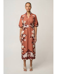 A woman modeling a rust-colored midi dress with a white and dark floral print, featuring short sleeves, a collar, and a matching fabric tie belt, set against a plain light gray studio background.