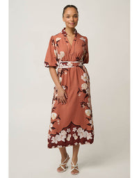 A woman modeling a rust-colored midi dress with a white and dark floral print, featuring a shawl collar, short sleeves, and a matching fabric tie belt, set against a plain light gray studio background.