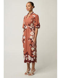 A woman modeling a short-sleeve midi dress with a floral print in terracotta, brown, and white, paired with white strappy heels, against a neutral studio background.