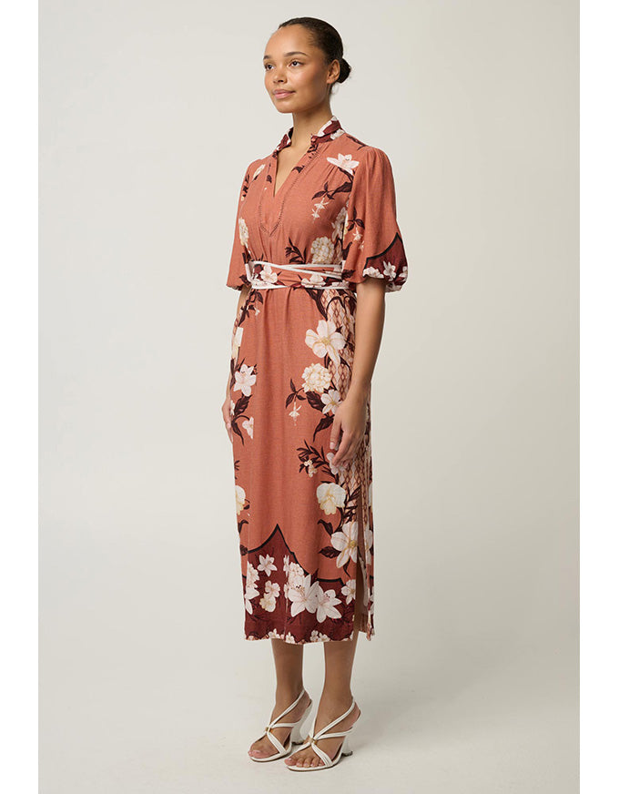 A woman modeling a short-sleeve midi dress with a floral print in terracotta, brown, and white, paired with white strappy heels, against a neutral studio background.