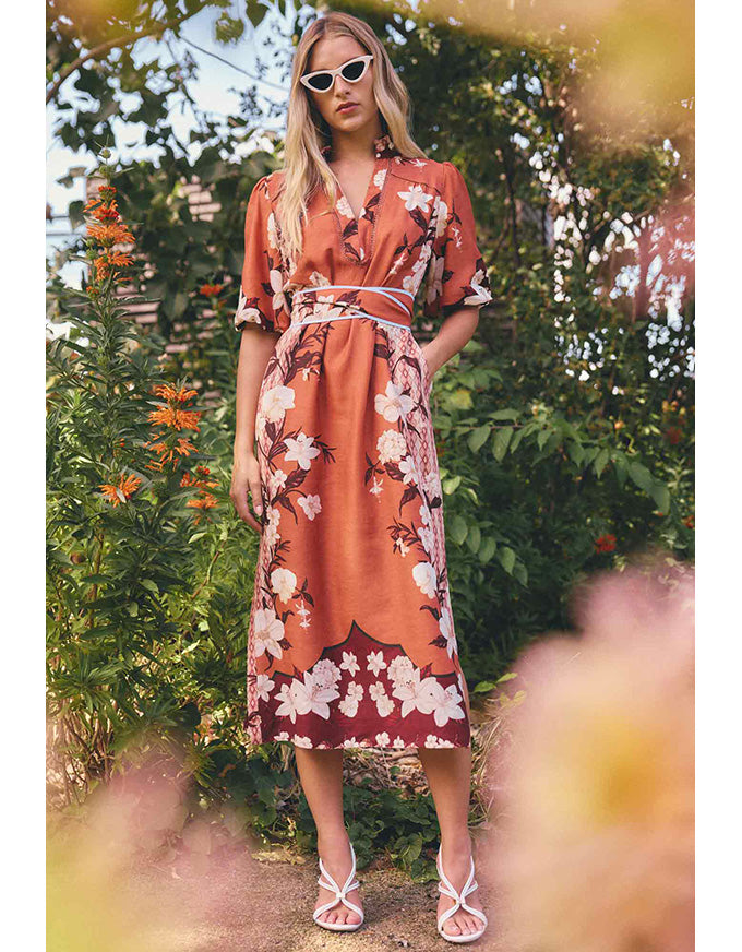 A woman wearing a rust-orange midi dress with a white floral print, a white tie belt, and white strappy sandals, standing in a garden setting with blurred foliage.