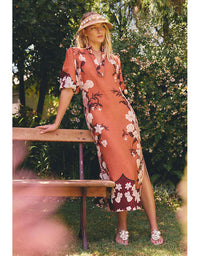 A woman stands outdoors on grass beside a wooden bench, wearing a peach-orange midi dress with a white and dark floral print, a matching hat, and sandals. The background is a lush green garden.