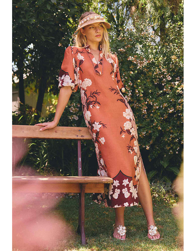 A woman stands outdoors on grass beside a wooden bench, wearing a peach-orange midi dress with a white and dark floral print, a matching hat, and sandals. The background is a lush green garden.