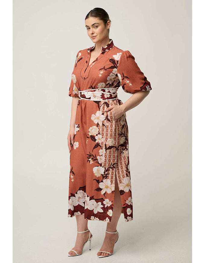 A woman modeling a rust-orange midi wrap dress with a white, black, and burgundy floral print, featuring a tie waist belt and short puff sleeves, set against a plain light gray studio background.