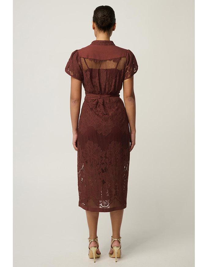 A woman viewed from the back wearing a rust-brown lace midi dress with short puff sleeves, sheer mesh back panels, and a matching fabric tie belt, paired with beige high-heeled sandals against a light gray studio background.