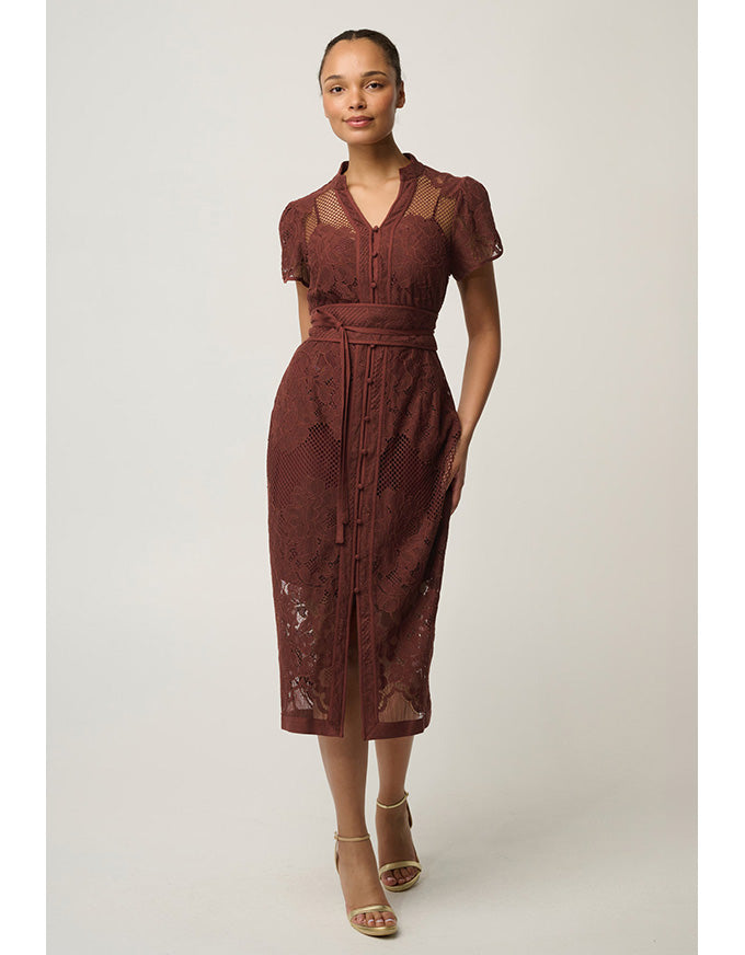 A woman modeling a rust-brown lace midi dress with sheer short sleeves, a V-neckline, and a matching waist tie, set against a plain light gray studio background.