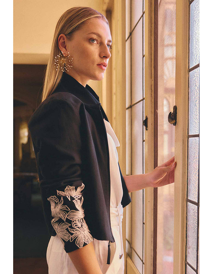 A woman in a black cropped jacket with white floral embroidery on the sleeve, worn over a white top, standing by a window.