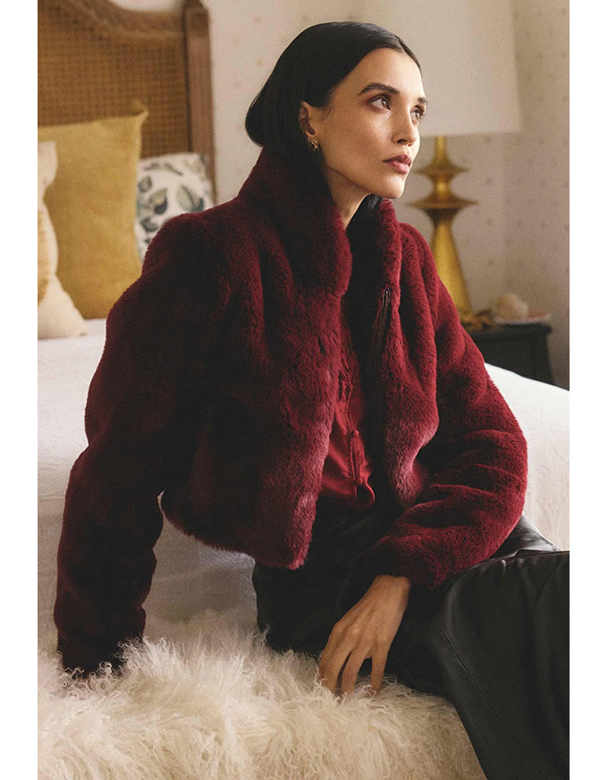 Woman wearing a red fur coat sitting on a bed in a bedroom setting.