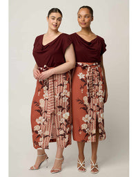 Two women modeling matching outfits with deep burgundy draped tops and rust-orange floral and patterned wrap-style bottoms, posed against a plain light beige studio background.