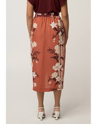 A woman wearing a rust-orange midi skirt with a white and dark floral print, paired with a dark red top and white heeled sandals, shown from the back against a light gray studio background.