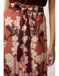 A close-up of a model wearing a rust-orange skirt with a white floral print, dark brown leafy stems, and a matching dark brown tie belt at the waist.
