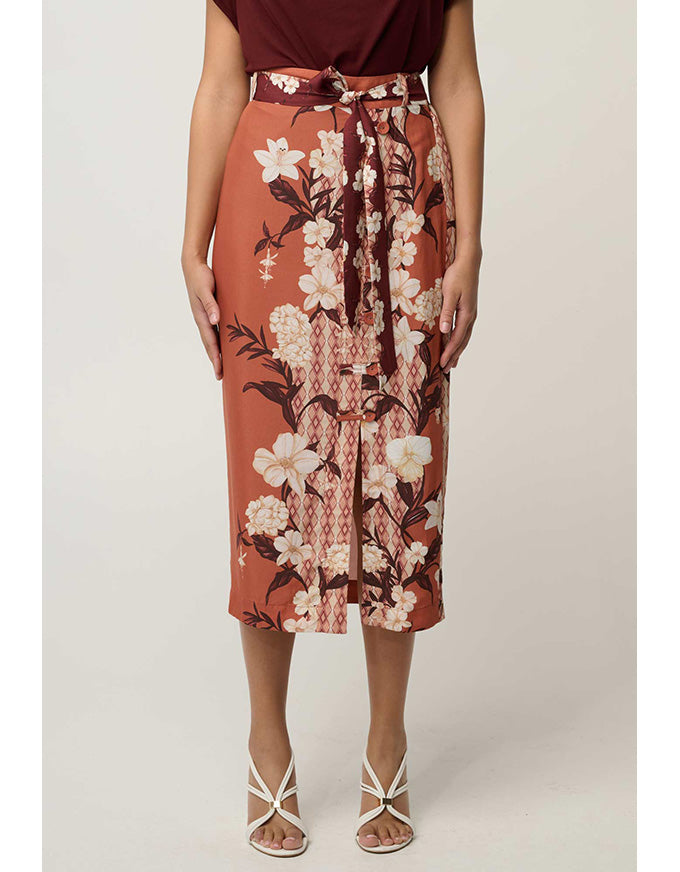 A model wearing a rust-orange midi skirt with a white and dark brown floral print, a matching tie belt, and white strappy heels against a light gray studio background.
