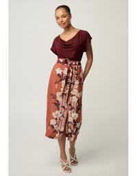 A woman modeling a two-piece outfit consisting of a dark burgundy cowl-neck short-sleeve top and a rust-orange midi skirt with a white and pink floral print and a tie waist.