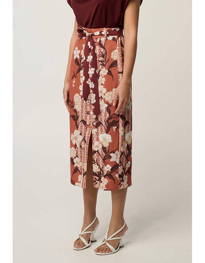 A model wearing a rust-orange midi skirt with a white and burgundy floral print, paired with a matching tie-waist belt and white strappy high-heeled sandals, set against a plain light beige studio background.