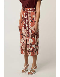 A model wearing a rust-orange midi skirt with a white and burgundy floral print, paired with a matching tie-waist belt and white strappy high-heeled sandals, set against a plain light beige studio background.