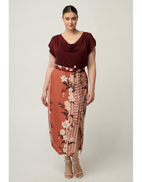 A woman modeling a two-piece outfit consisting of a dark burgundy cowl-neck short-sleeve top and a rust-orange wrap skirt with a floral print and patterned side panels, paired with metallic heeled sandals against a light gray studio background.
