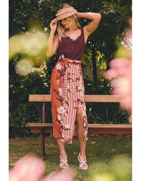A woman stands outdoors on a wooden bench in a garden, wearing a sleeveless dark burgundy top and a rust-orange midi skirt with a floral and geometric print, a matching belt, and a straw hat with flowers.