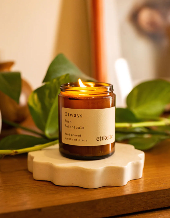 Candle in a glass jar with a label on a decorative coaster, surrounded by green leaves.