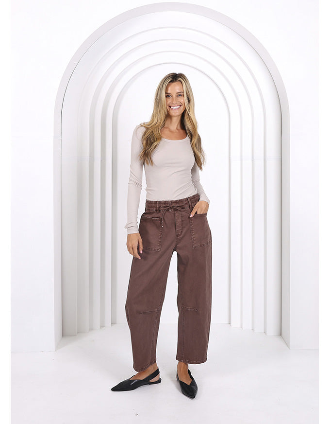 Woman wearing a beige top and brown pants standing in a white archway.