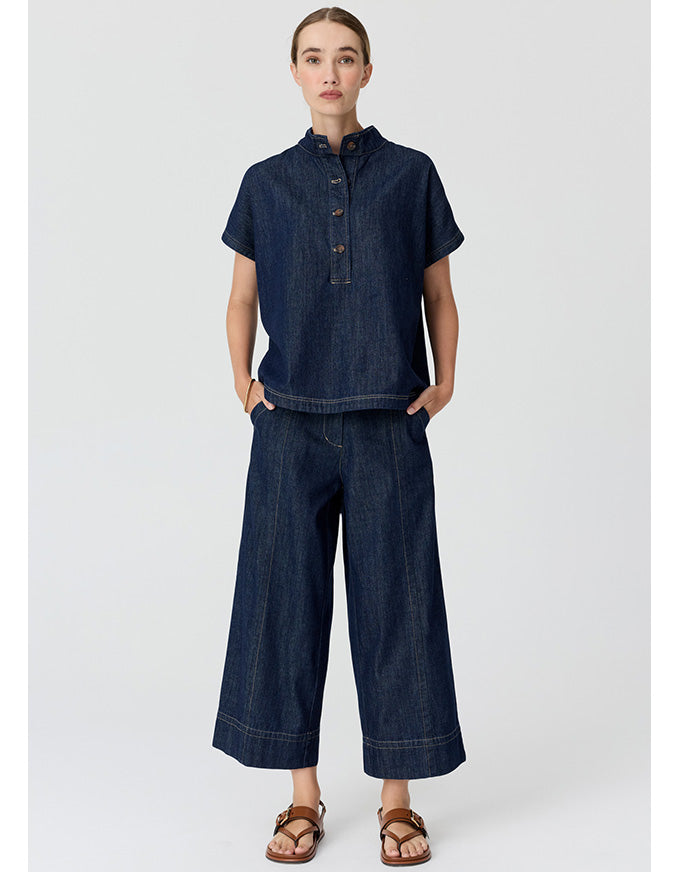 A model wearing a dark indigo denim two-piece set consisting of a short-sleeve button-up shirt and matching wide-leg cropped pants, paired with brown leather sandals, against a light gray studio background.