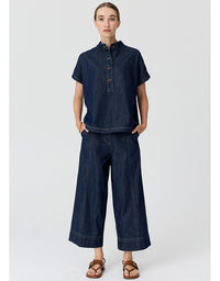 A model wearing a dark indigo denim two-piece set consisting of a short-sleeve button-up shirt and matching wide-leg cropped pants, paired with brown leather sandals, against a light gray studio background.