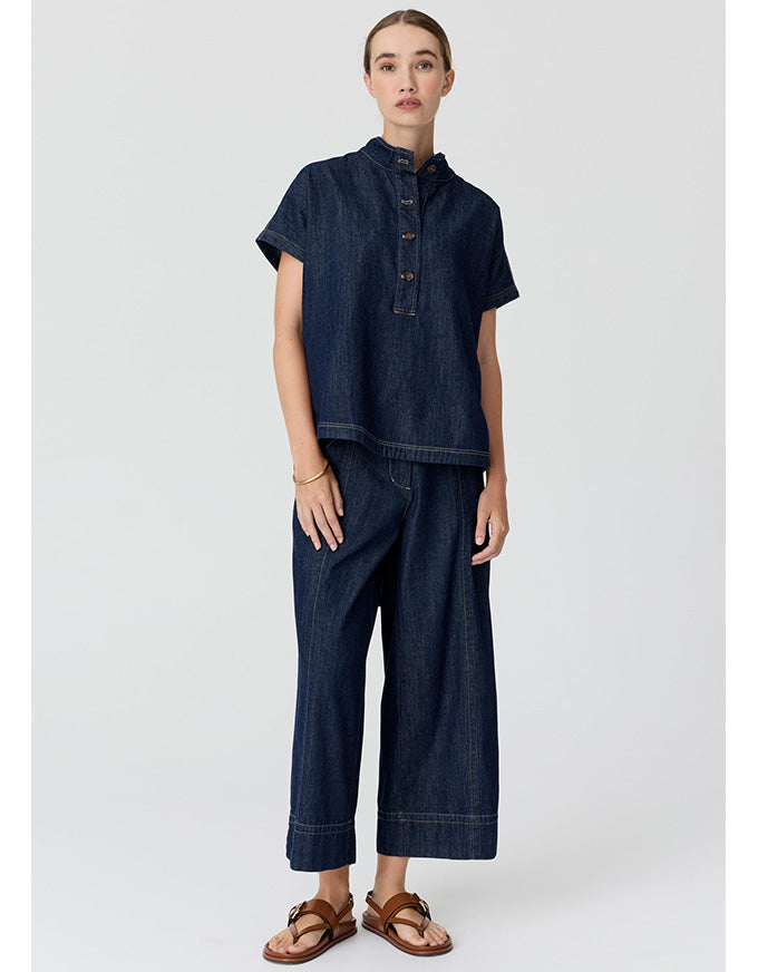 A model stands against a light gray studio background wearing a dark indigo denim two-piece set consisting of a short-sleeve button-front top and matching wide-leg cropped pants, paired with brown leather sandals.