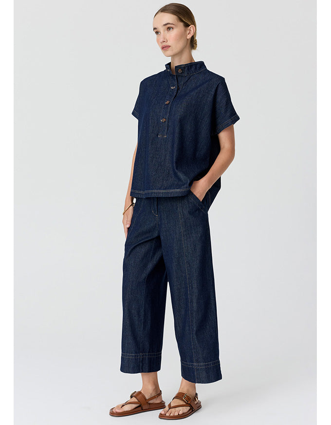 A woman modeling a dark indigo denim two-piece set consisting of a short-sleeve button-up shirt and matching wide-leg cropped pants, paired with brown leather sandals, against a light gray studio background.