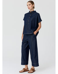 A woman modeling a dark indigo denim two-piece set consisting of a short-sleeve button-up shirt and matching wide-leg cropped pants, paired with brown leather sandals, against a light gray studio background.